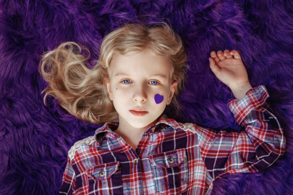 Pretty blonde girl child lying on purple fluffy rug with purple violet felt paper heart on cheek