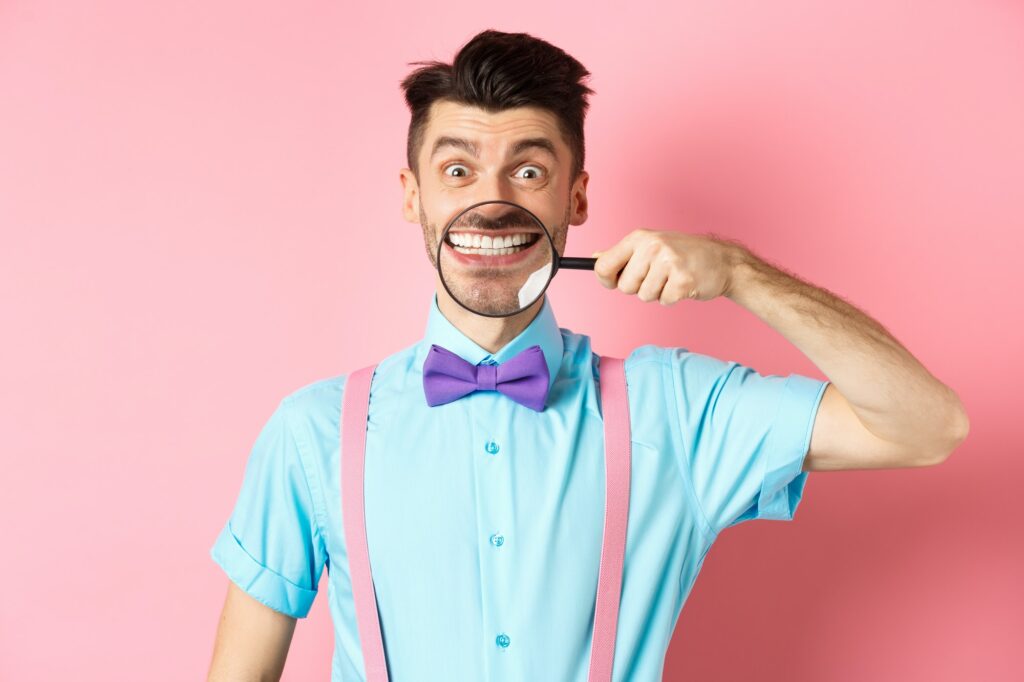 Funny caucasian guy in bow-tie showing his white smile teeth with magnifying glass, looking cheerful