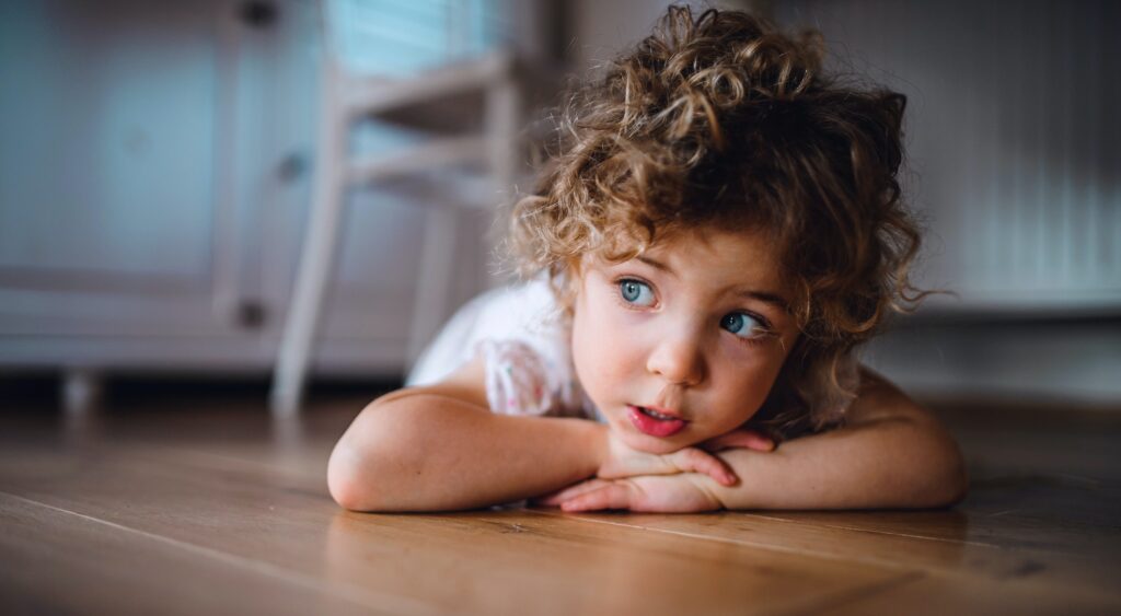Portrait of cute small girl lying on floor under table indoors at home, looking away