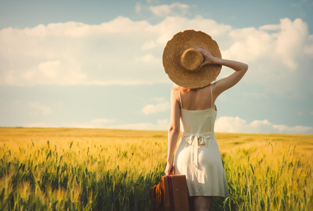 Blode woman in hat with suitcase in wheat field in sunset time