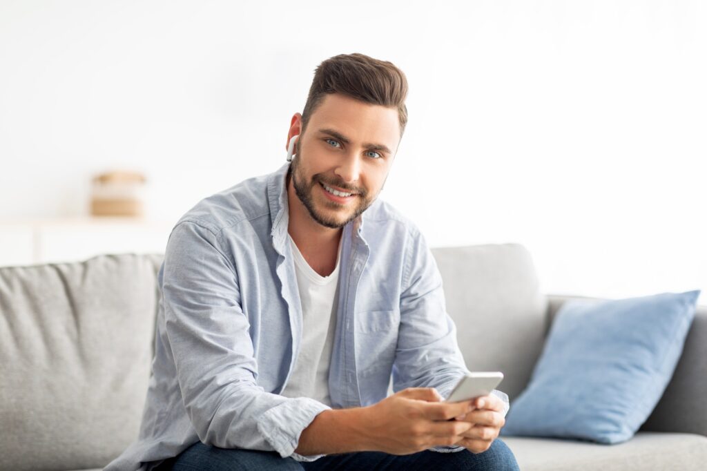 Handsome millennial man in earbuds using smartphone and smiling to camera, sitting on sofa at home