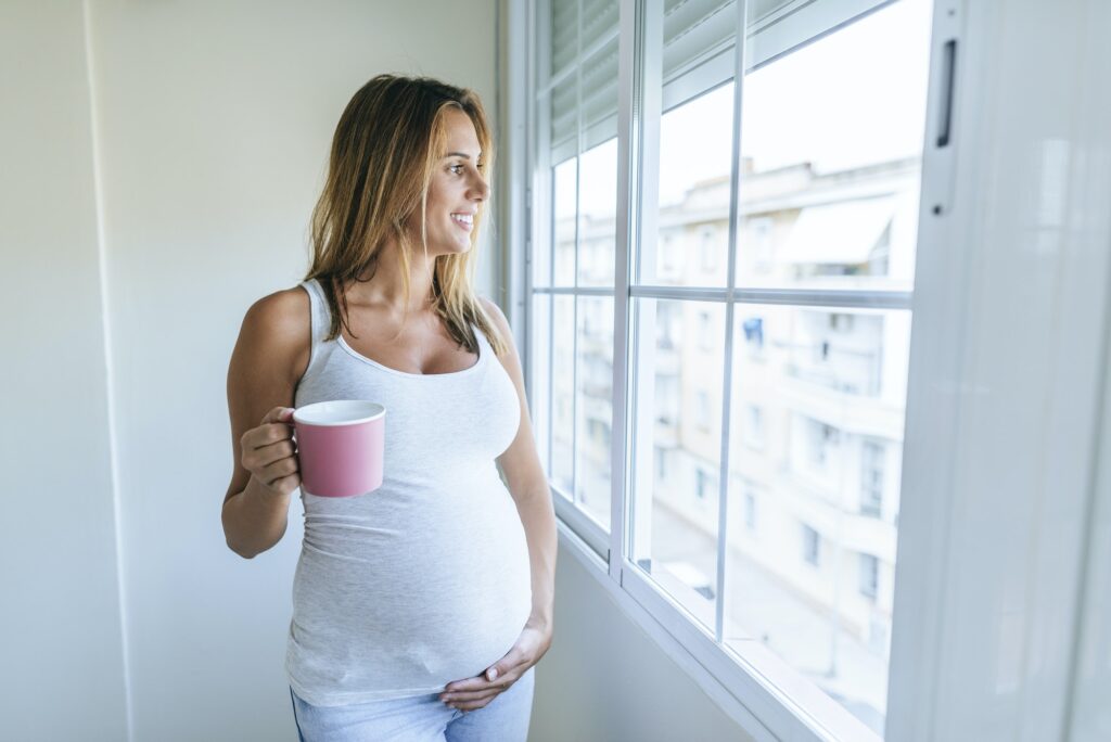 Pregnant woman looking out of window holding mug