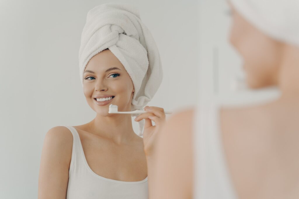Beautiful young woman with healthy perfect smile brushing teeth and looking in mirror