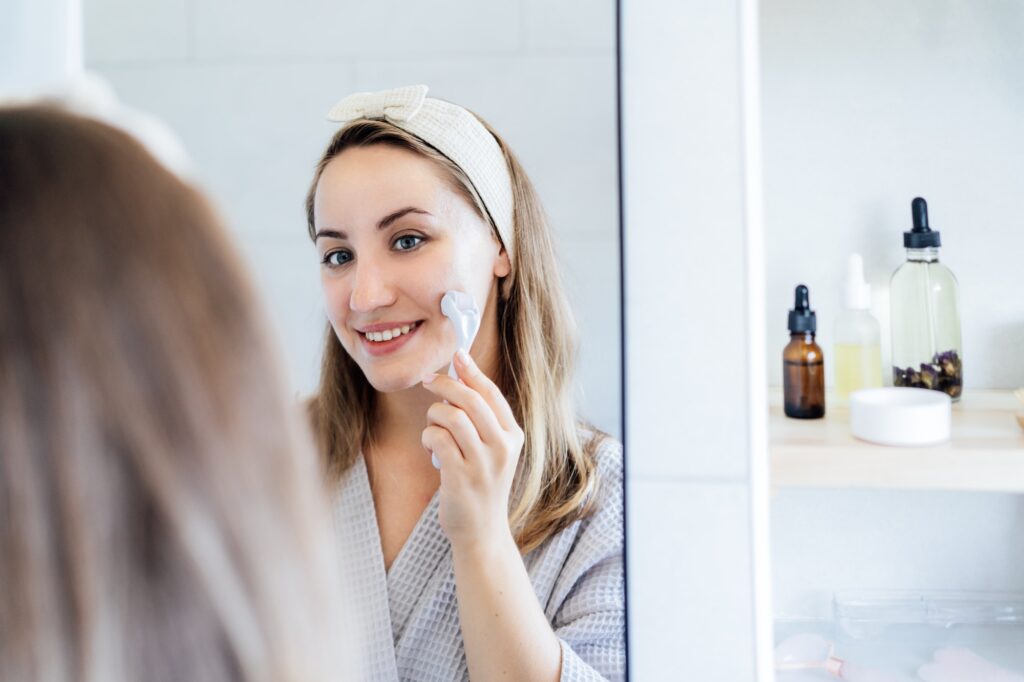 Young woman in bathrobe looking in mirror and making face massage with microneedle meso derma Roller