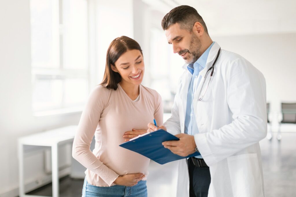 Male doctor showing clipboard with medical test results to happy pregnant lady during meeting in