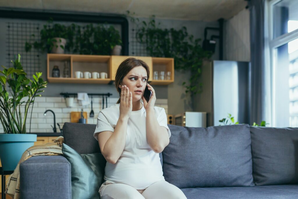 Sad pregnant woman at home anxious talking on the phone sitting on the couch